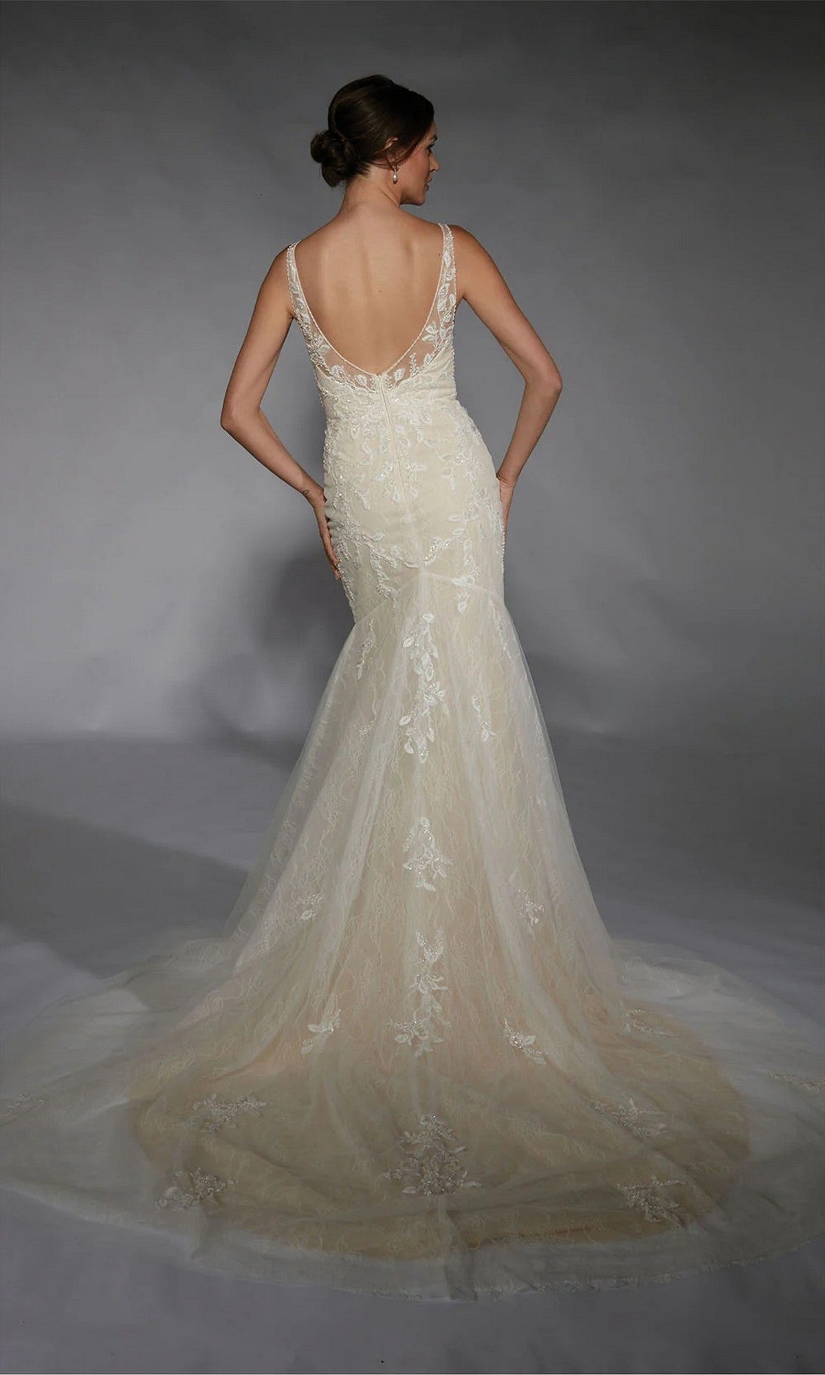 A woman faces away, wearing the Alyce 7119 Long White Dress with a mermaid silhouette, beaded lace, and a sheer train embellished with floral embroidery, set against a plain gray background.